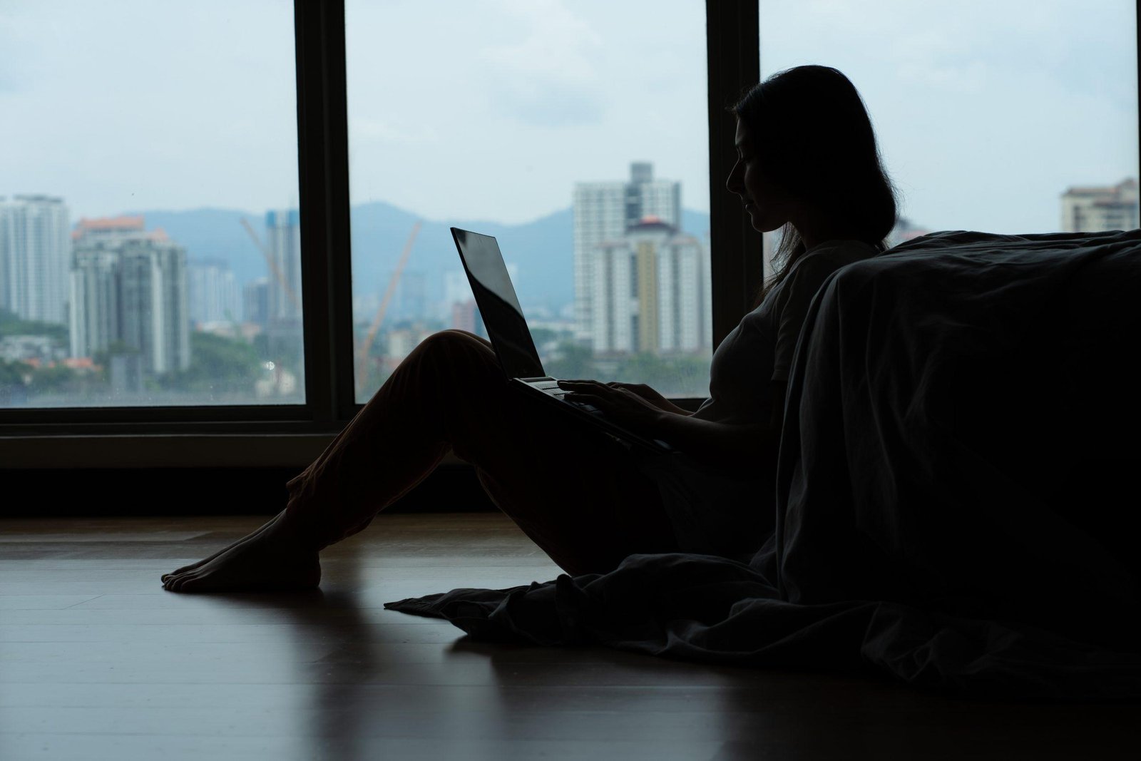 Siluet girl working on a laptop and drinking coffee, sitting on the floor near the bed by the panoramic window with a beautiful view from the high floor. A cozy workplace. Shopping on the Internet.