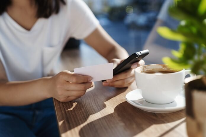 Close-up of women hands leaning on coffee table, holding mobile phone and plastic credit card. Girl paying online with smartphone, sending money for cappuccino
